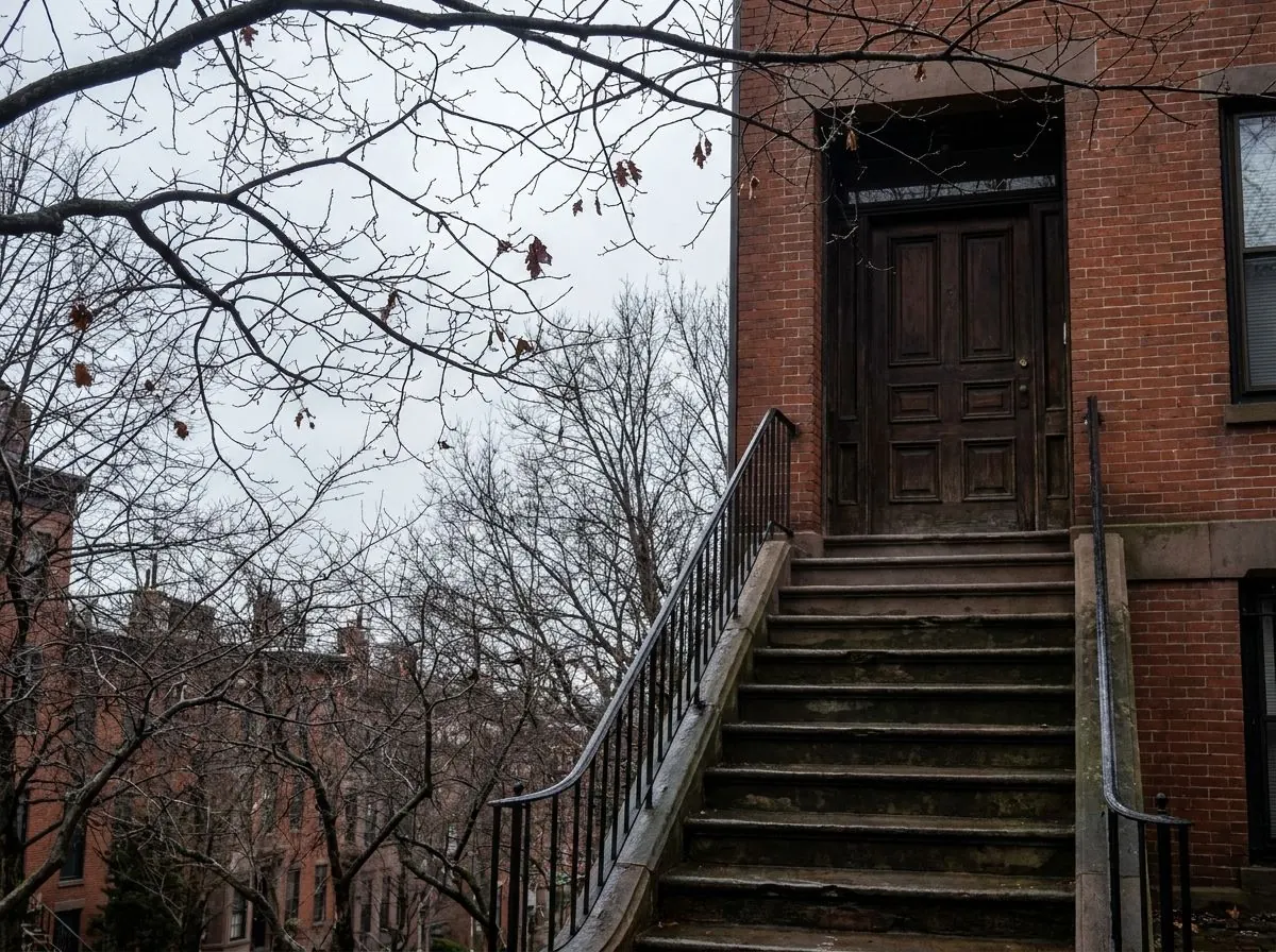 A narrow brick row house entrance reached by a steep flight of stone steps, with a wrought iron handrail and a heavy panelled front door.