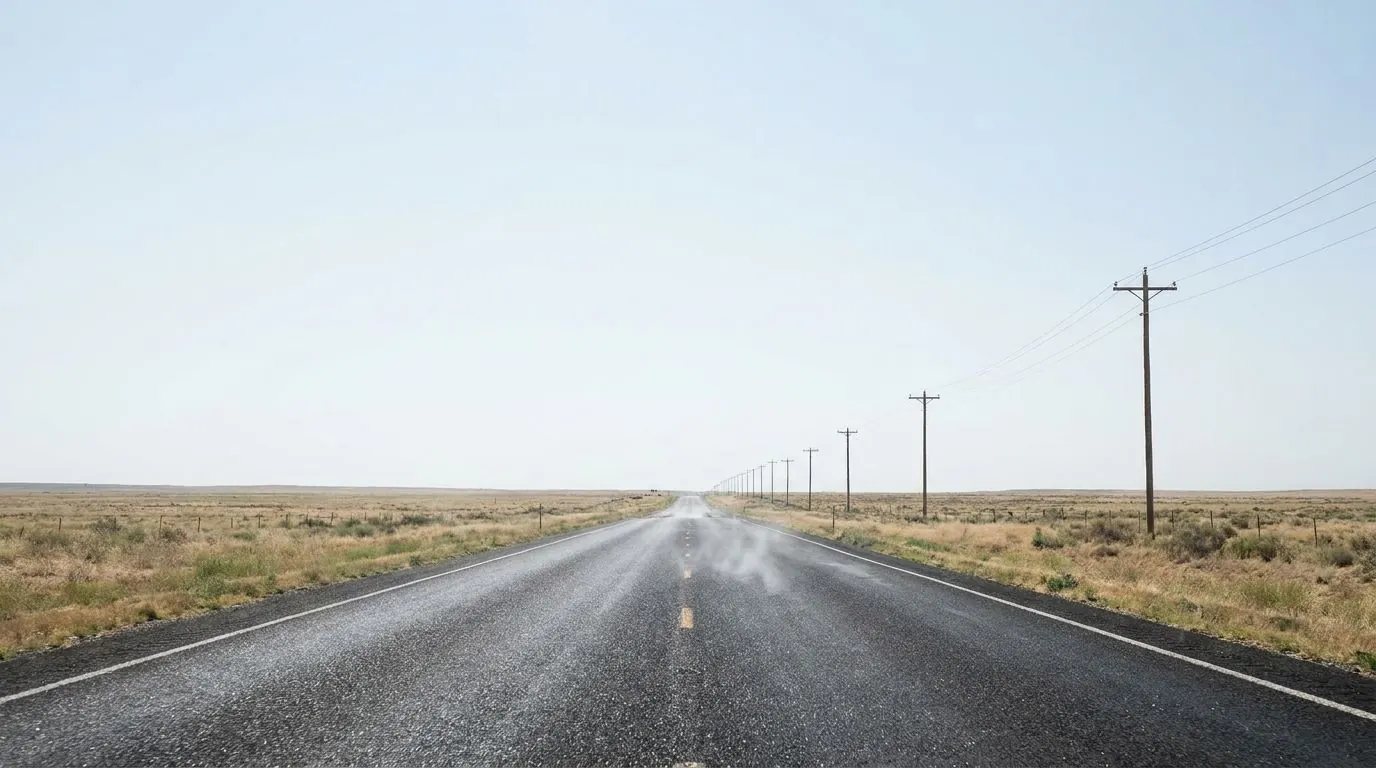 A straight two-lane highway running across flat open grassland toward a distant horizon under an enormous pale sky.
